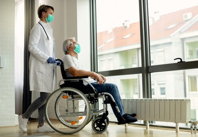 Female doctor pushing senior man in wheelchair to the window during coronavirus lockdown.